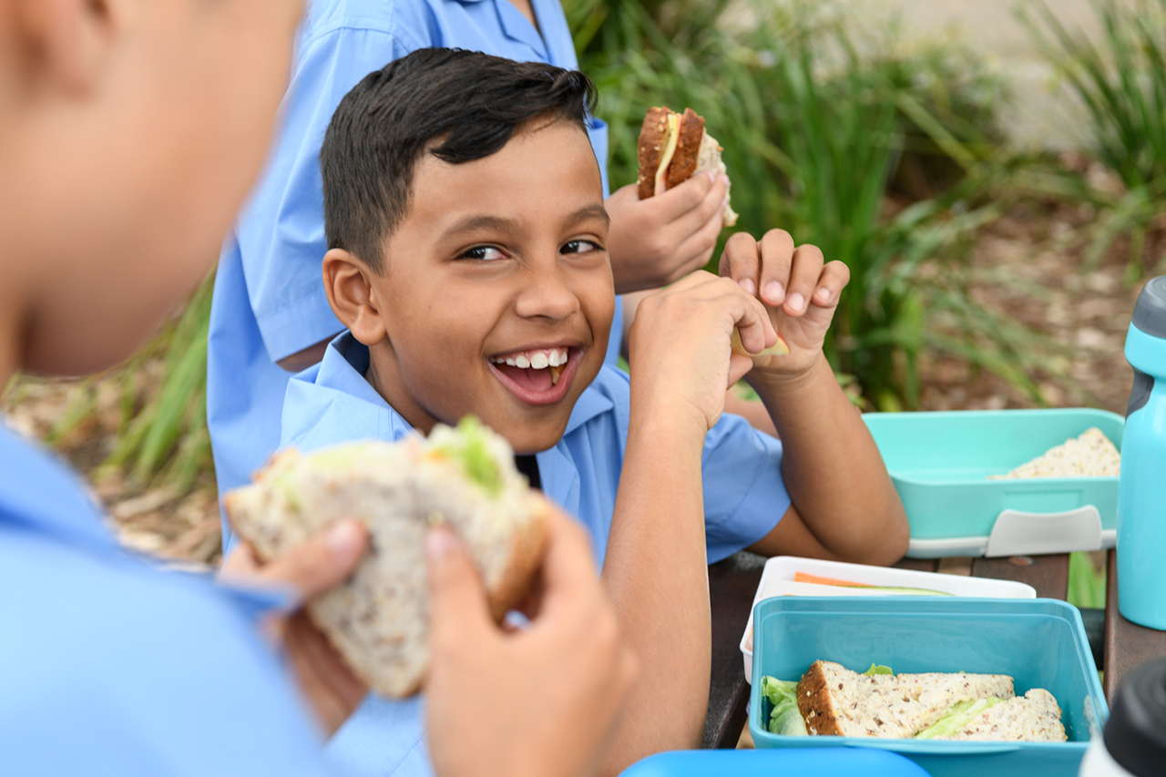 Young school boy in blue shirt smiling and looking away with lunch box on table