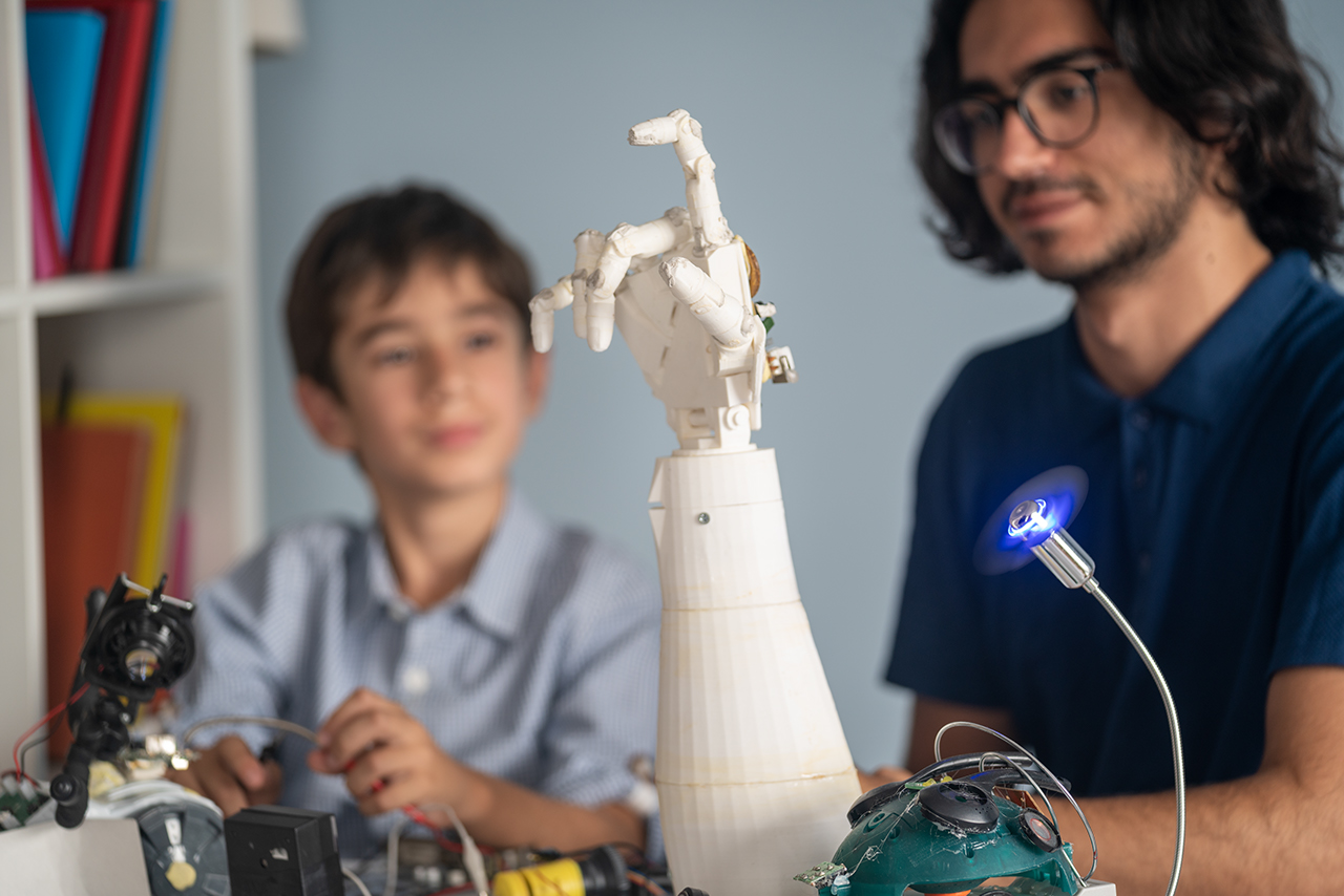 College student teaching student coding and robotics. A robotic hand is seen on desk.