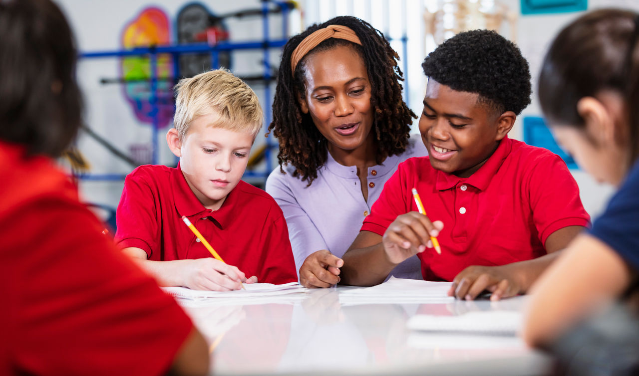 An African-American teacher helping a multiracial group of middle school students in class. The students are sitting at a table writing in notebooks, doing a writing assignment or taking a test.